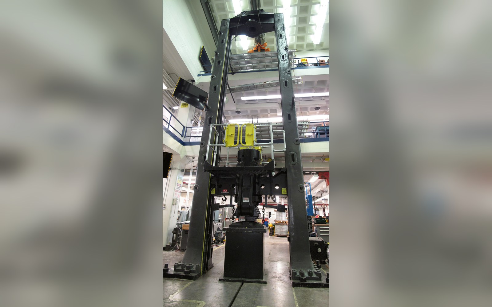 A tall industrial press frame stands on a factory floor surrounded by mezzanine walkways